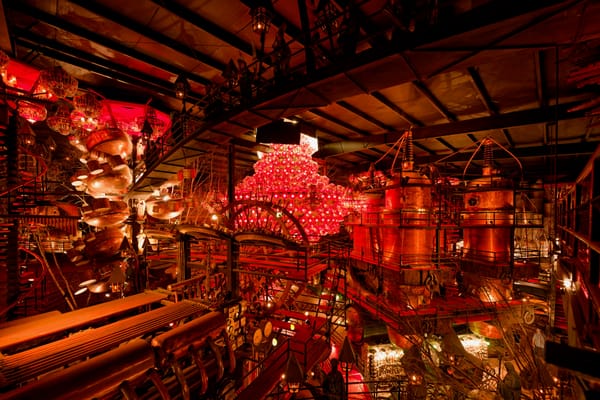 A view of the Organ Room, a chaotic assortment of metal machinery, glass lamps, and metal catwalks covered in carpet, all housed in a very large multi-story hangar.